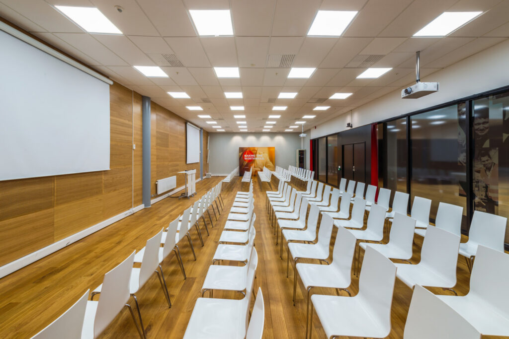 Konferenssal med rader av vita stolar under ett jämnt rutnät av energieffektiva takarmaturer som ger klart, bländfritt arbetsljus perfekt för presentationer och events, professionell belysning från PROLJUS AB