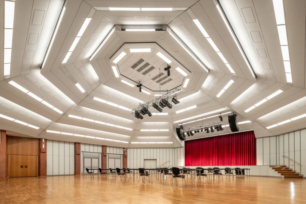 Storslagen auditoriumbild med flerskiktat hexagonalt tak där integrerade linjära LED‑armaturer och upphängda scenstrålkastare ger jämn, energieffektiv och flexibel belysning som visar PROLJUS ABs skräddarsydda ljuslösningar för professionella arenor