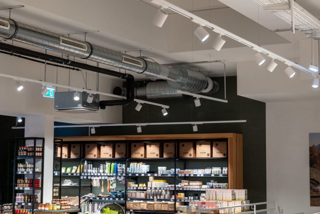 Butiksinteriör med exponerade ventilationskanaler i taket och vita skenspottar monterade i skenor som riktar varmt, fokuserat ljus över hyllor och varor, energieffektiv och säljdrivande belysningslösning från PROLJUS AB som framhäver produkter och skapar inbjudande köpupplevelse