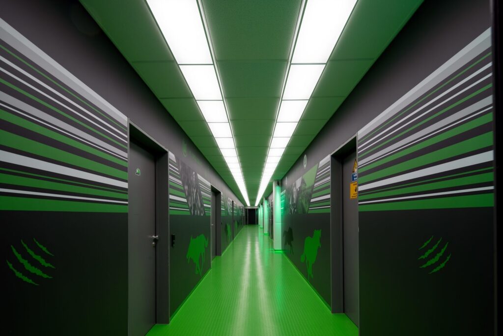 Lång korridor belyst av linjära, energieffektiva takpaneler från PROLJUS AB som ger jämnt, klart vitt ljus och framhäver lokalens gröna accenter och moderna industridesign