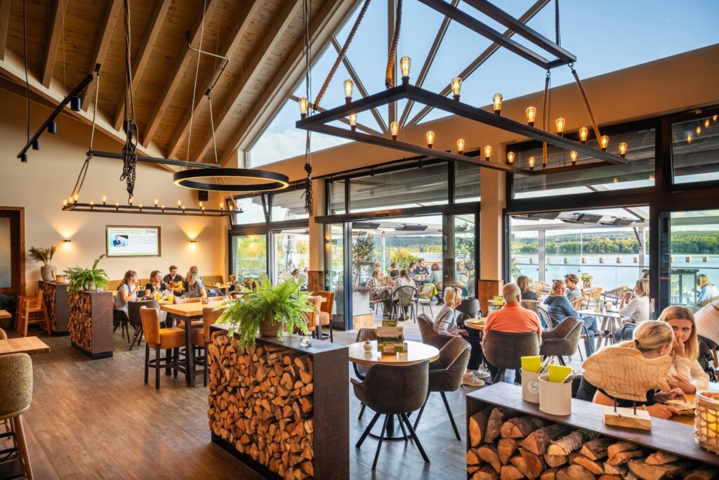 Modern restaurang med sjöutsikt – ljus matsal och terrass, matgäster njuter av mat och dryck, rustik träinredning.