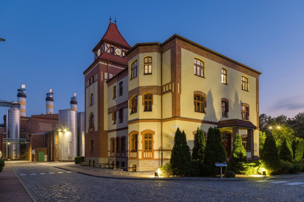 Kvällsbild av en upplyst tegelbyggnad och industrimiljö där PROLJUS ABs armaturer levererar varma uplights som framhäver fasaddetaljer, spotlights som lyser upp träd och gångvägar och dekorativa gatlyktor som skapar stjärnklara effekter och visar PROLJUS ABs energieffektiva lösningar