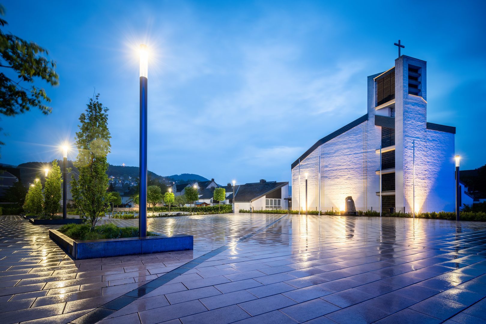 Kvällsbild av upplyst torg och kyrka där PROLJUS AB:s moderna, energieffektiva ljusarmaturer kastar varmt vitt och kallt blått sken över blanka plattor och framhäver arkitekturen, en attraktiv lösning för stadsrum och försäljning