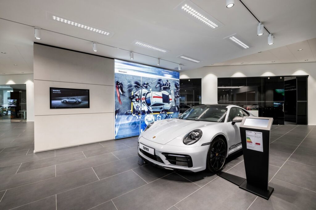 Modern bilsalong med vit sportbil i förgrunden och PROLJUS AB:s belysning i fokus, breda infällda LED-paneler i taket för jämnt allmänljus, riktbara skenspotlights som accentuerar bilen och infällda downlights längs väggarna som framhäver skyltning, energieffektiva armaturer med hög färgåtergivning som skapar en premiumatmosfär och ökar produktens attraktionskraft