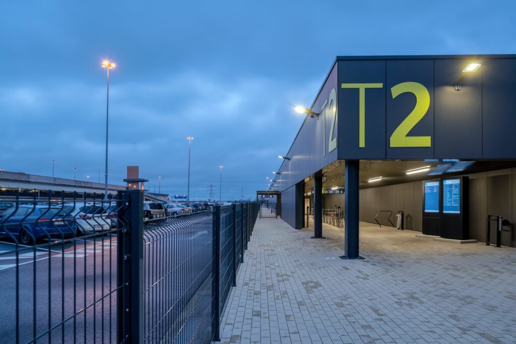 Terminal T2 i skymning upplyst av moderna stolp- och väggarmaturer som kastar jämnt kallvitt ljus över gångväg och parkering och visar PROLJUS ABs energieffektiva, robusta belysningslösningar