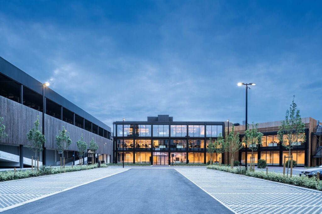 Kontorsbyggnad i skymning belyst av moderna master med PROLJUS AB:s luminärer som sprider jämnt, varmt och energieffektivt ljus över parkeringsytor och entré, elegant och robust belysning för kommersiella miljöer