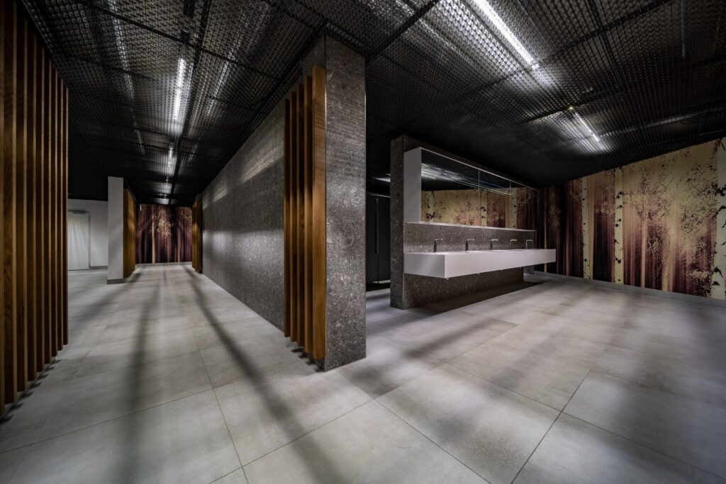 Modernt offentligt utrymme upplyst av PROLJUS AB:s slimmade LED-linjer monterade i svart nätat tak som skapar jämn, riktad belysning och dramatiska skuggor över klinkergolv och handfatsområde, energieffektiva och eleganta luminärer perfekta för kommersiella miljöer