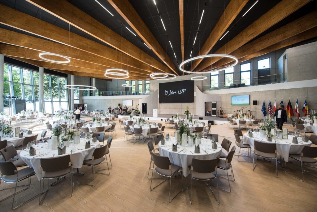 Stor ljus bankettsal med flera hängande cirkelformade LED-armaturer i olika storlekar och höjder monterade längs träbjälkar, som skapar varm jämn allmänbelysning och elegant atmosfär, från PROLJUS AB energismarta dimbara armaturer idealiska för konferenser event och hotell