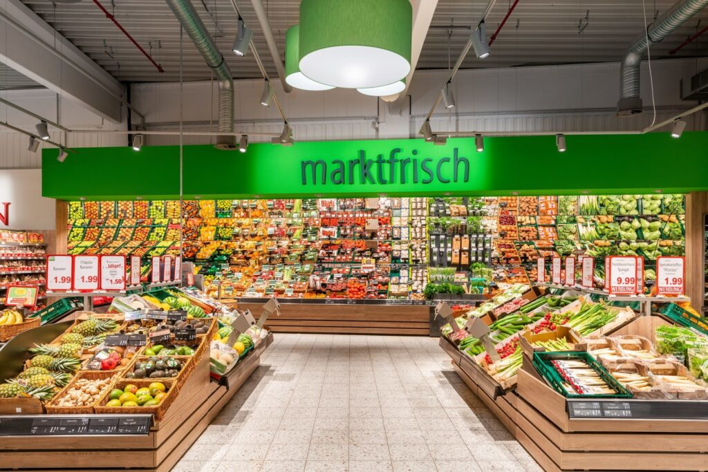 Interiör i mataffär med stora gröna cylinderformade pendellampor och riktade spotlights som skapar varmt, jämnt ljus över frukt och grönsaker, effektiv butikbelysning från PROLJUS AB som lyfter produktpresentation och ökar försäljningen