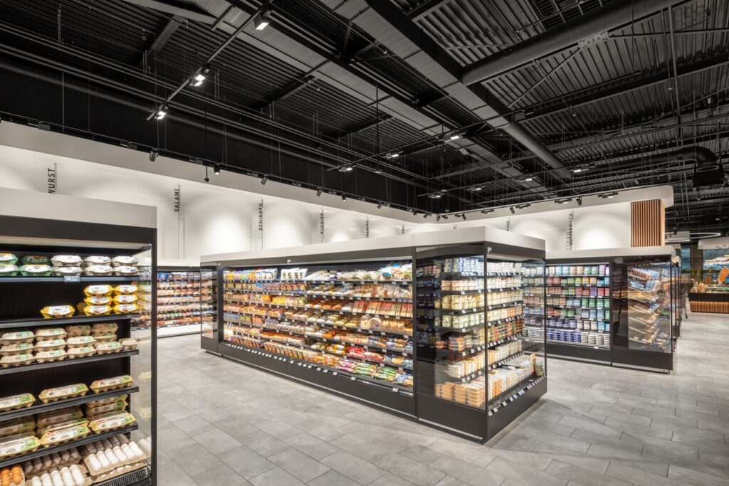 Invändig butiksmiljö med svart öppet undertak och pendlande linjära LED-armaturer samt riktbara spottar som ger klar, jämn och färgkorrigerad belysning på hyllor och varor — PROLJUS ABs energieffektiva, justerbara takarmaturer optimerar exponering och minskar driftkostnader