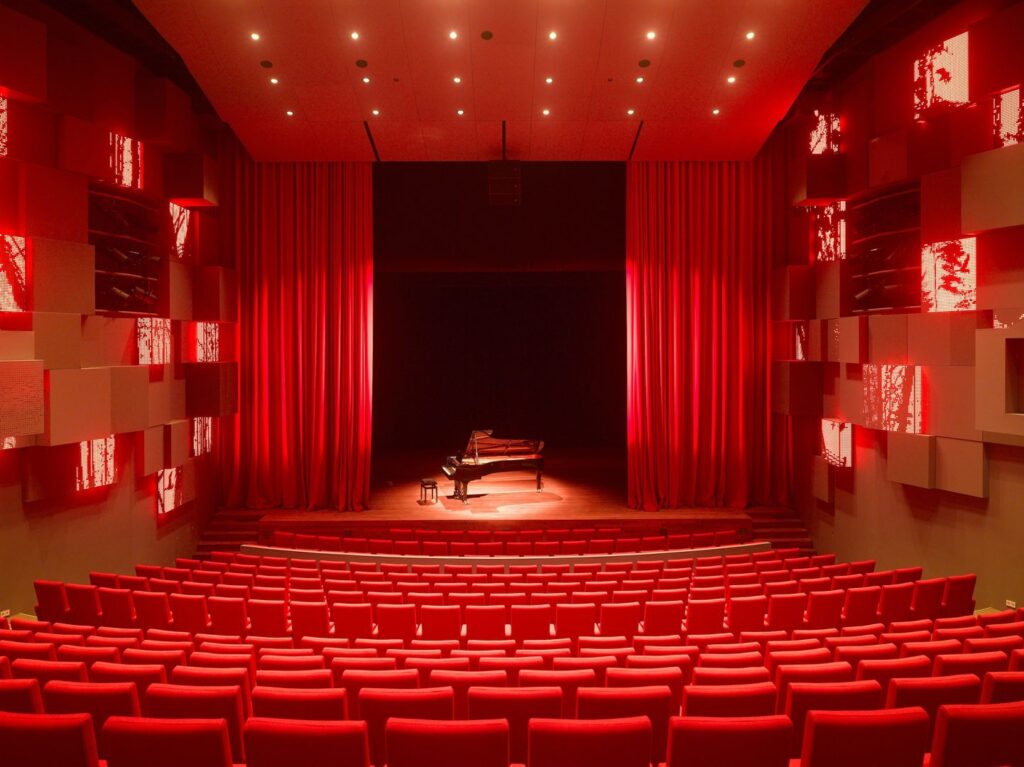 Teatersal med rader av röda stolar och en flygel på scenen upplyst i dramatiskt rött av diskreta takspotlights och väggmonterade LED-armaturer levererade av PROLJUS AB för publikmiljöer