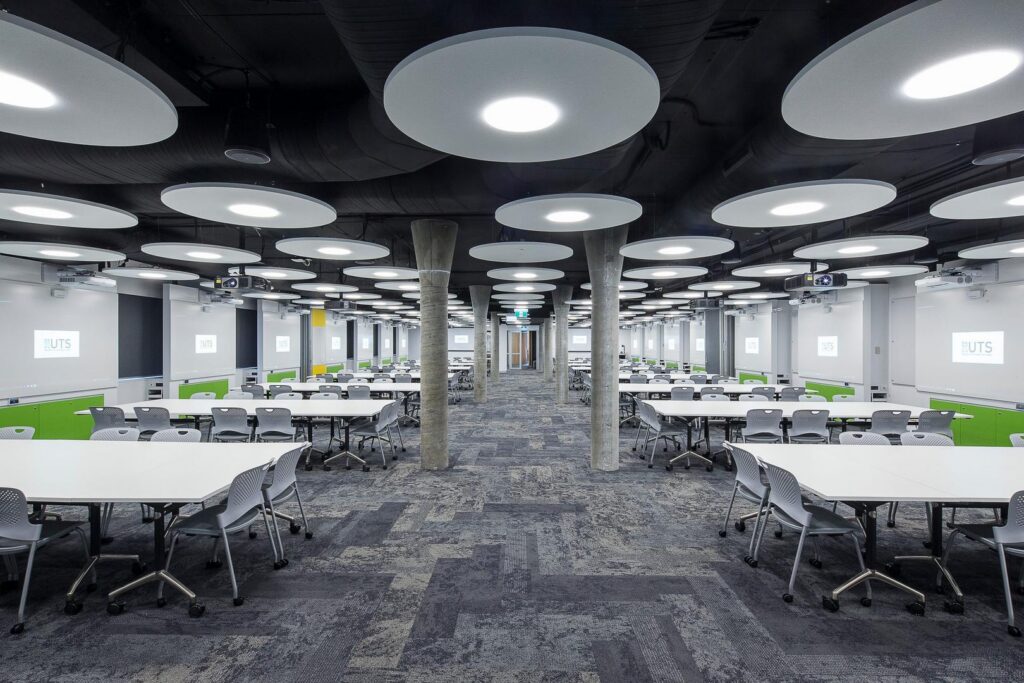 Stort modernt undervisningsrum med rader av stora runda takarmaturer som ger jämnt bländfritt ljus över bord och stolar, en energieffektiv och designad belysningslösning från PROLJUS AB som lyfter pedagogiska och konferensmiljöer