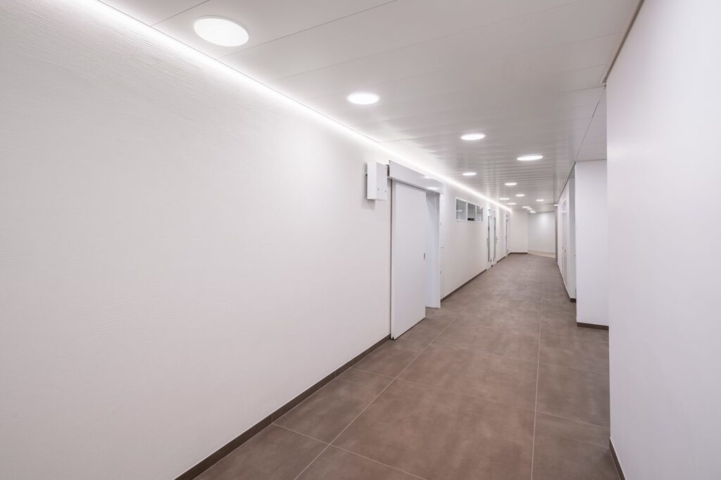 Lång vit korridor upplyst av runda infällda LED-armaturer i taket och en subtil LED-slinga längs taklisten från PROLJUS AB som ger jämn, energieffektiv och behaglig belysning för moderna kontor och offentliga miljöer