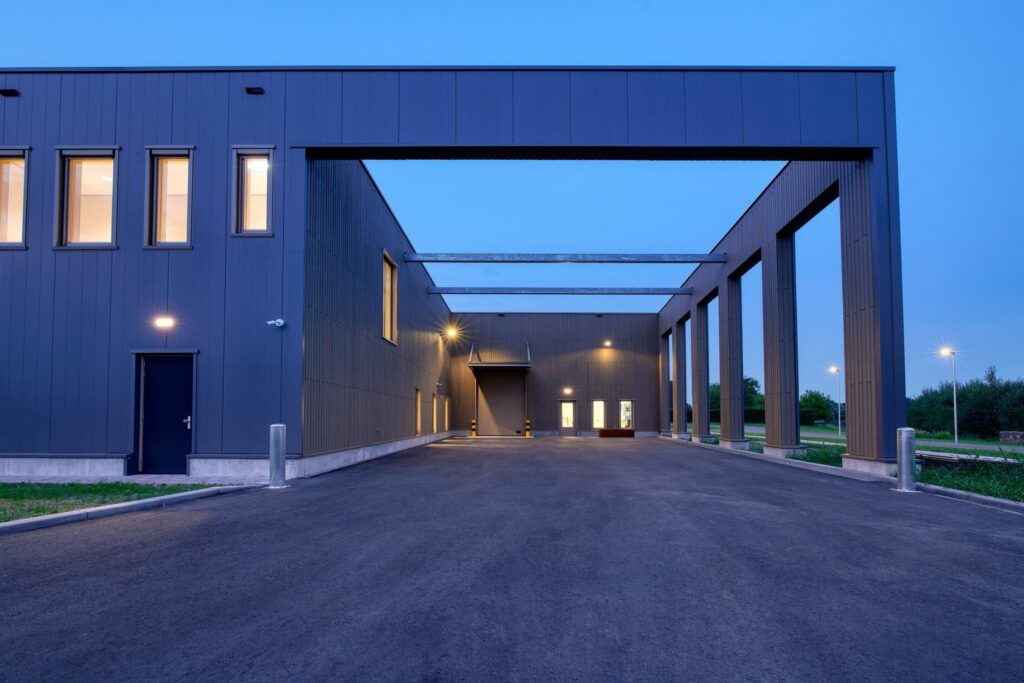 Kvällsbild av modern industrifasad upplyst av PROLJUS AB:s väggarmaturer, pollare och stolpbelysningar som ger jämn, energieffektiv och hållbar ljusspridning över infarten och det öppna atriet, perfekt för att öka säkerhet och visuellt intryck