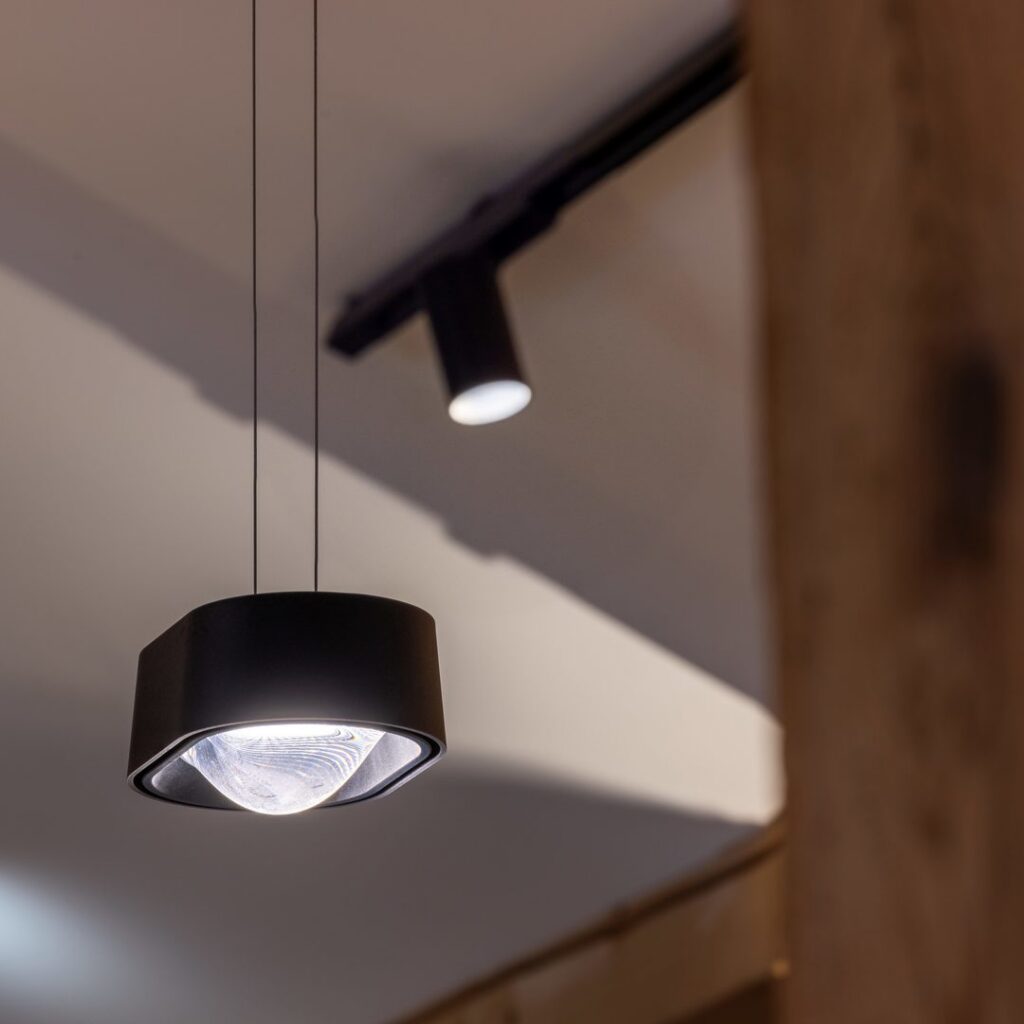 Detaljbild av en svart rund pendelarmatur med tydlig lins och en riktbar spot på takskena i varmt vitt ljus, elegant och energieffektiv ljuslösning från PROLJUS AB för butiker och moderna miljöer