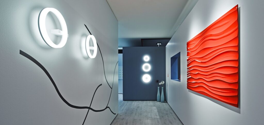 Visningsrum med stilrena ringformade LED-väggarmaturer i olika storlekar som avger kallt vitt sken mot ljusa väggar, tre cirkulära ringlampor monterade vertikalt på en mörk fondvägg och accentbelysning som framhäver en röd vågformad väggpanel, energieffektiva och designade armaturer från PROLJUS AB perfekta för moderna hem och offentliga miljöer