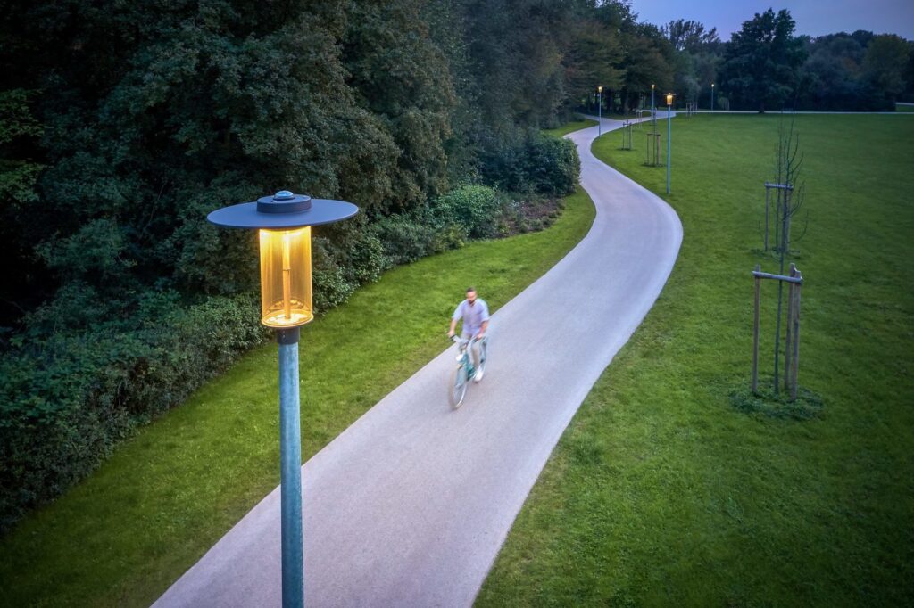 Parkvy med slingrande gång- och cykelväg upplyst av PROLJUS ABs moderna armaturer, närbild på en stilren lyktstolpe i galvaniserat stål med varm, energieffektiv LED som skapar trygghet och atmosfär längs stigen och flera identiska armaturer i perspektiv visar en enhetlig och hållbar lösning för offentliga miljöer