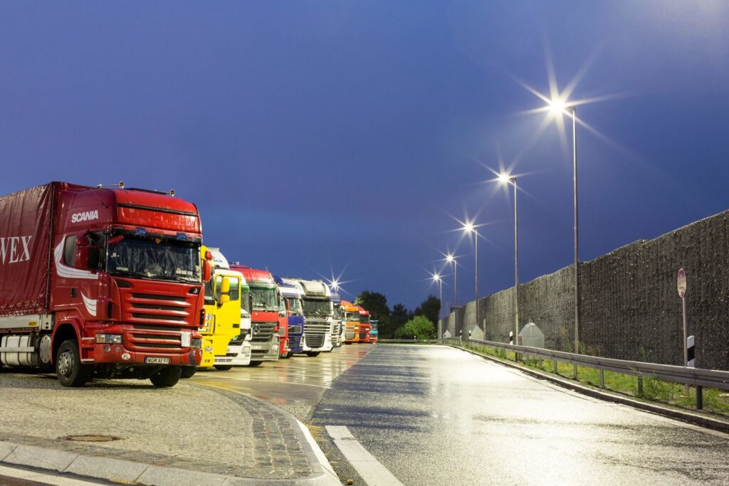 Rad av moderna, ljusstarka gatlyktor med stjärnformade ljussken över blanka vägytor och parkerade lastbilar, en tydlig presentation av PROLJUS AB:s energieffektiva och driftssäkra vägbelysningsarmaturer som ökar säkerhet och minskar kostnader