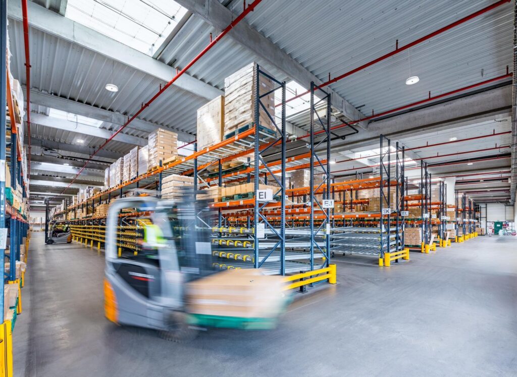 Stor lagerhall upplyst av jämnt fördelade takarmaturer och LED-highbay-armaturer monterade i takbalkar, kompletterade med takljus som ger dagsljusinsläpp, energieffektiv och bländfri industribelysning som förbättrar säkerhet och produktivitet, idealisk lösning från PROLJUS AB