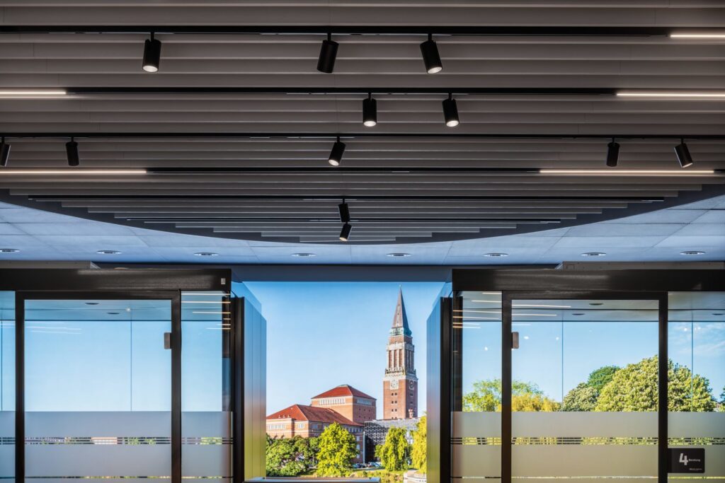 Takmonterade svarta riktbara cylinderformade spotlights och linjära LED-skenor integrerade i ett lamelltak som skapar jämnt energieffektivt arbetsljus över glaspartier i en modern entré, elegant och funktionell belysningslösning från PROLJUS AB