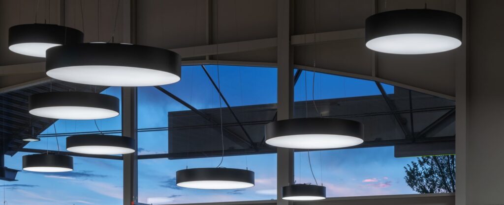 Flera cirkulära svarta takpendlar med jämnt diffust vitt LED-ljus hängande i olika höjder framför stora fönster i skymning, modern och energieffektiv belysning från PROLJUS AB idealisk för kontor och offentliga miljöer