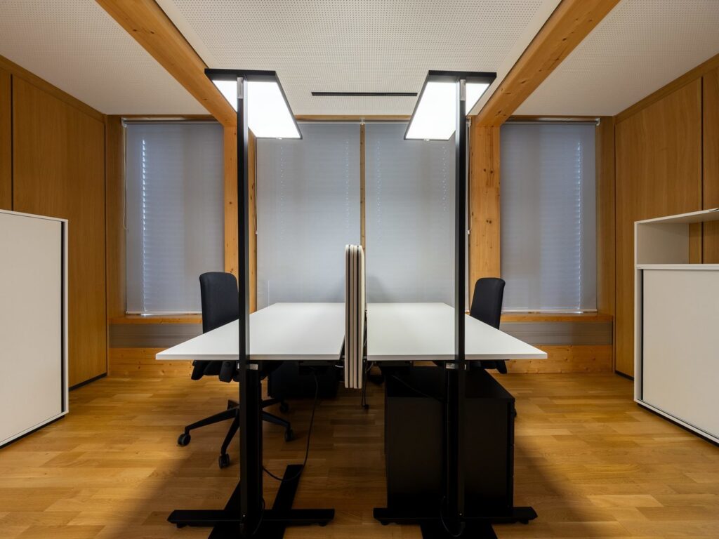 Två rektangulära LED-armaturer från PROLJUS AB monterade på svarta stolpar över skrivbord, ger jämnt och bländfritt arbetsljus i en varm träklädd kontorsmiljö och kombinerar stilren design med funktion för ökad produktivitet