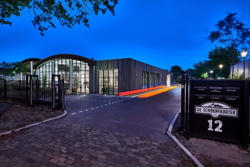 Kvällsbild av modern företagsbyggnad och infart upplyst av PROLJUS AB:s armaturer, pollare och fasadbelysning som framhäver glasfasaderna, skapar trygg väl upplyst infart med energieffektiva varmvita LED-lösningar och inbjudande atmosfär för kunder