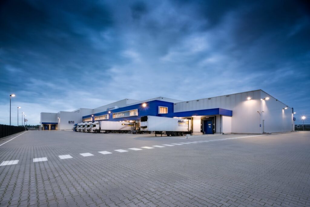 Exteriör kvällsbild av ett modernt lager med lastkajer belysta av energieffektiva väggarmaturer och mastbelysning från PROLJUS AB som ger jämnt, kraftfullt LED-ljus över ytorna och framhäver säkerhet, lägre driftkostnader och modern estetik för industribyggnader