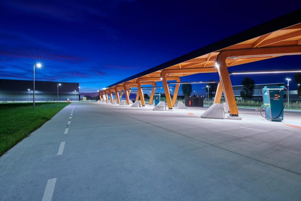 Nattbild av en EV‑laddstation upplyst av PROLJUS AB:s moderna, energieffektiva armaturer med starka stolpbelysningar och långa LED‑linjer under träbågar som skapar jämn, trygg och visuellt attraktiv belysning perfekt för kommersiella anläggningar