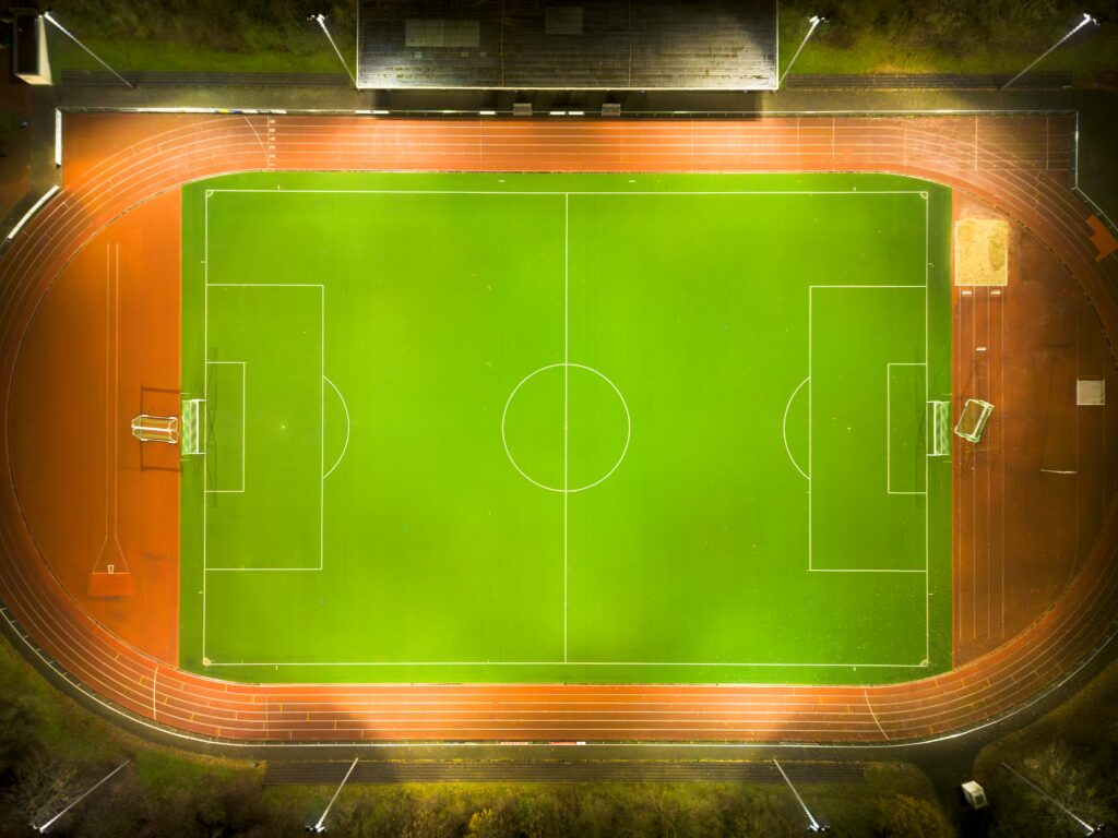 Fågelperspektiv över en kvällsupplyst fotbollsplan och löparbana där PROLJUS AB:s kraftfulla, energieffektiva LED-armaturer runt arenans kant ger jämn, riktad belysning som förbättrar synlighet, säkerhet och minskar driftkostnaderna