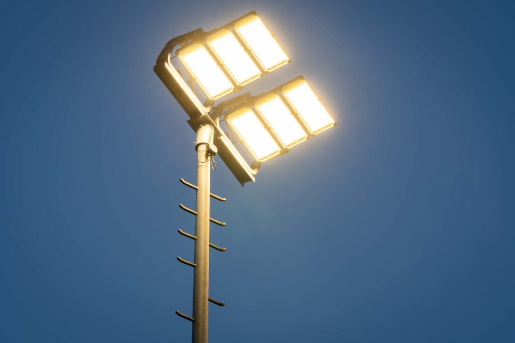 Hög stolpe med två rader om tre kraftfulla rektangulära LED-armaturer som lyser varmvit mot blå kvällshimmel, robust industri- och idrottsbelysning designad och levererad av PROLJUS AB