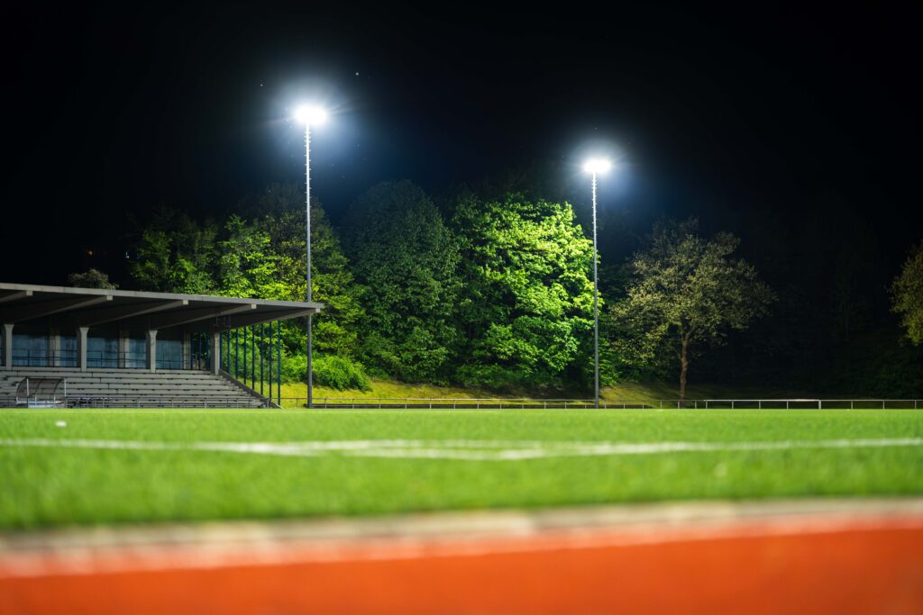 Nattbild av en upplyst idrottsplats med två höga PROLJUS AB:s strålkastare som sprider starkt och jämnt vitt ljus över spelplanen och läktaren och framhäver träd i bakgrunden perfekt för tryggt spel och evenemang