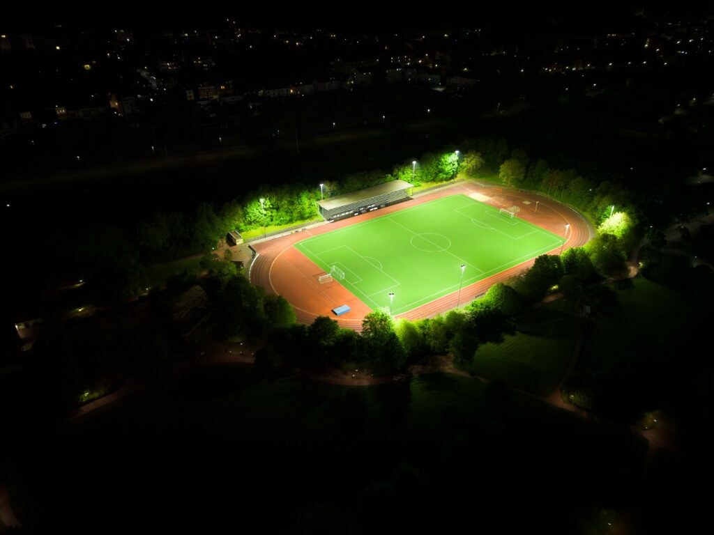 Luftbild på en upplyst fotbollsplan med löparbana och läktare omgiven av träd, jämnt och bländfritt belyst av energieffektiva LED-armaturer från PROLJUS AB som framhäver spelplanens färger och säkerställer optimal sikt för kvällsaktiviteter