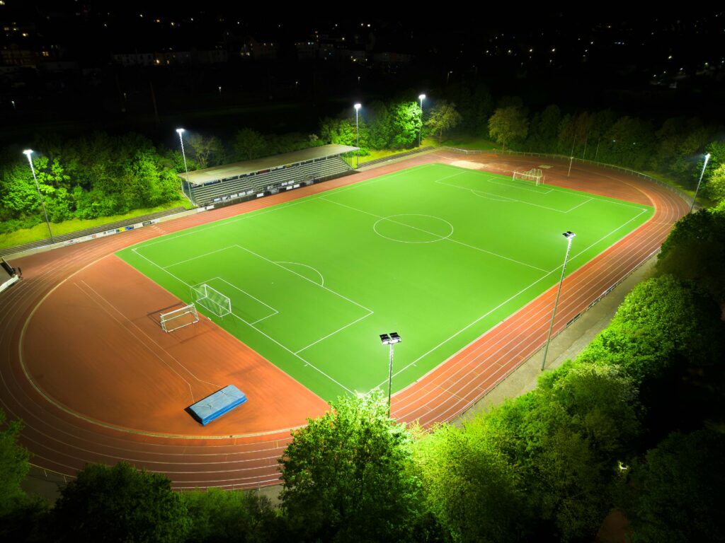 Flygfoto av en upplyst fotbollsplan på natten där höga master med moderna LED-armaturer från PROLJUS AB sprider jämnt, bländfritt och energieffektivt ljus över spelplan och löparbana och förbättrar säkerhet och publikupplevelse