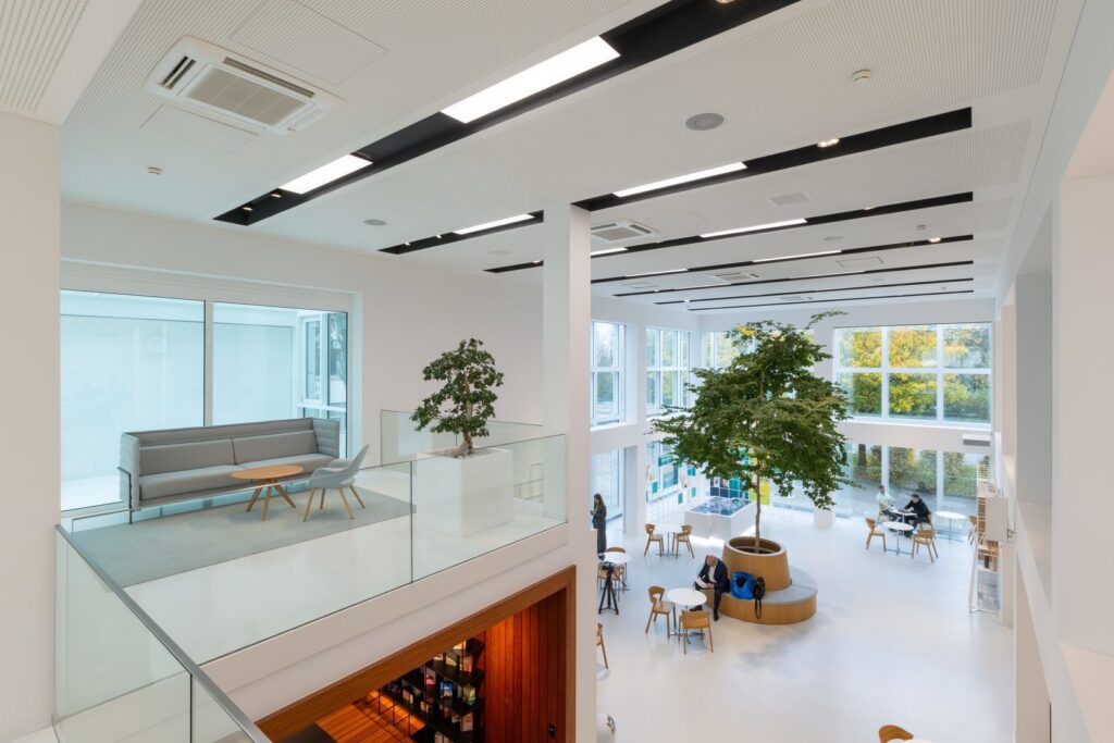 Öppet tvåvåningskontor med vita perforerade innertak där svarta infällda kanaler visar PROLJUS ABs energieffektiva linjära armaturer och spotlights som ger ett jämnt dagsljusliknande sken över växter och mötesytor, idealiskt för arkitektoniska och kommersiella projekt