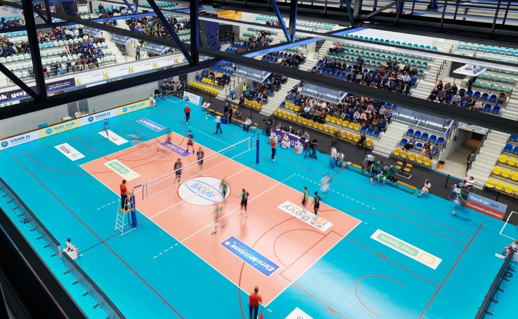 Vy uppifrån över en inomhusarena där robusta LED-armaturer monterade på stålfackverk ger jämn, bländfri och kraftfull belysning över volleybollplanen som visar PROLJUS AB:s energieffektiva, långlivade och lättinstallerade belysningslösningar som förbättrar sikt, TV-bild och driftsekonomi för sportarenor