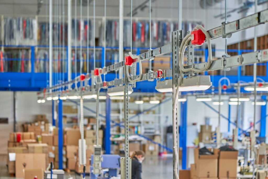 Närbild på hängande industriluminärer och linjära LED-armaturer monterade i räls med kabelkanaler och röda kopplingar i ett lager, klart vitt ljus som jämnt belyser arbetsytor, robust och energieffektiv lösning från PROLJUS AB