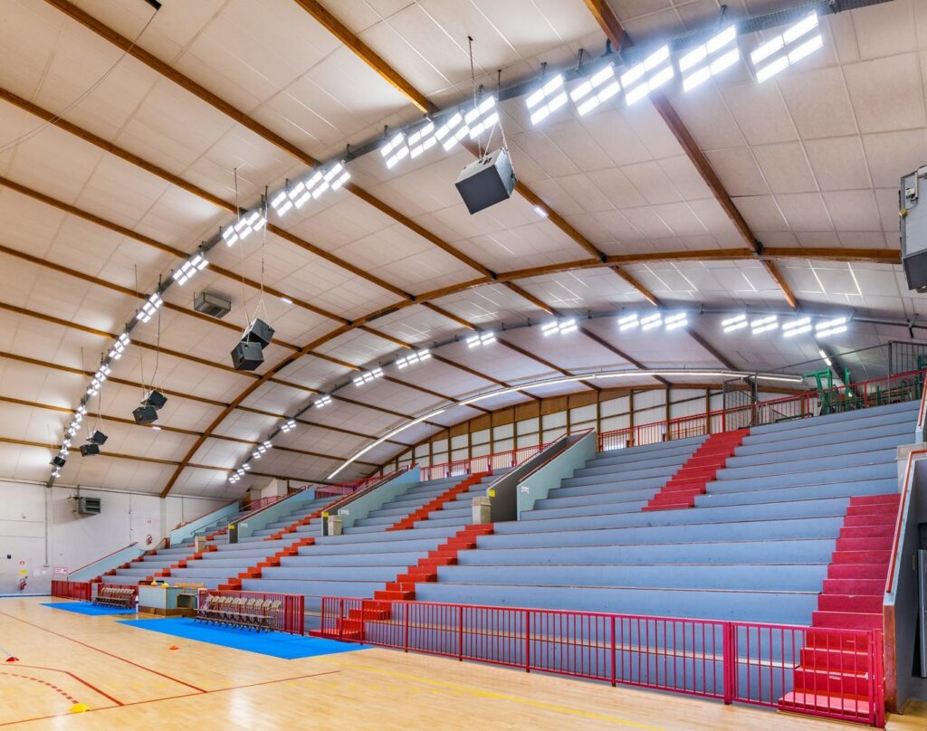 Ljust sporthallstak med radvis monterade energisnåla LED-armaturer och hängande ljusmoduler som levererar jämn högintensiv belysning över läktare och spelplan levererade och installerade av PROLJUS AB