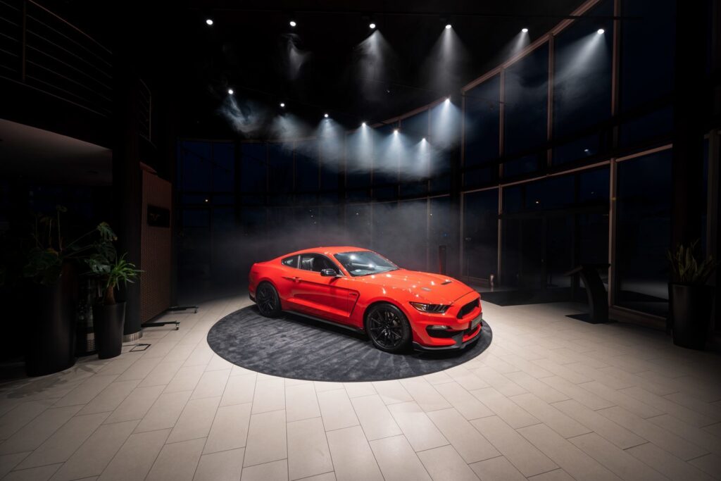 Showroom med en röd sportbil upplyst av PROLJUS ABs professionella spotlights som skapar skarpa ljusstrålar och dramatisk rökig atmosfär för säljande produktpresentation