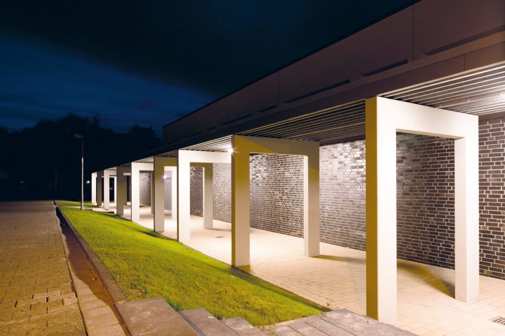 Nattbild av täckta entrégångar upplysta av PROLJUS AB:s varmvita, energieffektiva armaturer som ger jämn, dekorativ och säker belysning mot tegelväggarna och framhäver byggnadens moderna formspråk, idealisk för utomhusprojekt