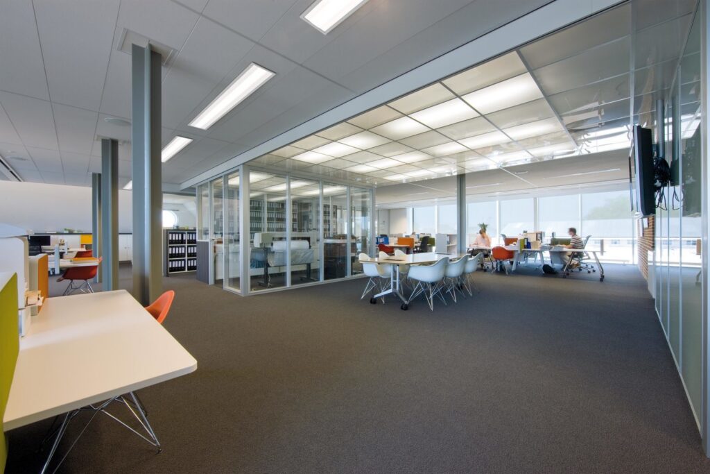 Öppet kontorslandskap med stora jämnt lysande takpaneler i rutmönster över glasad mötescell och rektangulära infällda armaturer längs taket som ger bländfri arbetsbelysning, jämn ljusfördelning och energieffektiv drift från PROLJUS AB