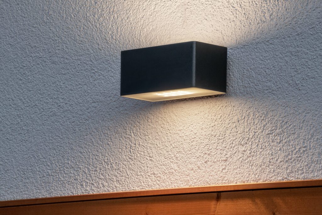 Modern svart LED-vägglampa med upp- och nedljus på vit putsad vägg ovanför träpanel.