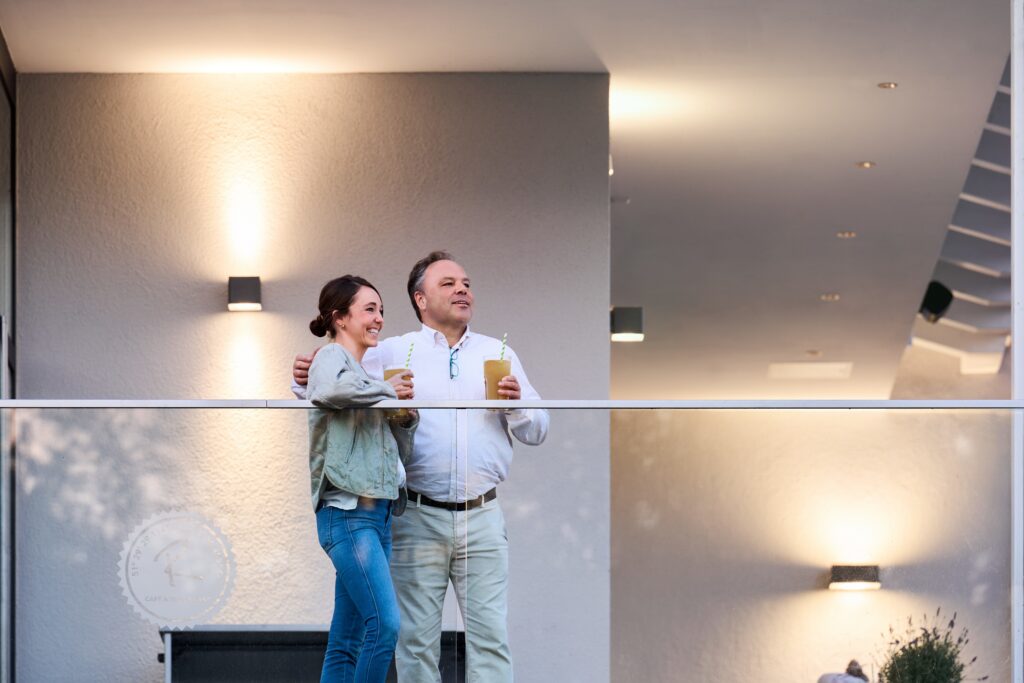 Två personer njuter av drinkar på modern balkong i kvällsljus – avslappnad uteplats med glasräcke