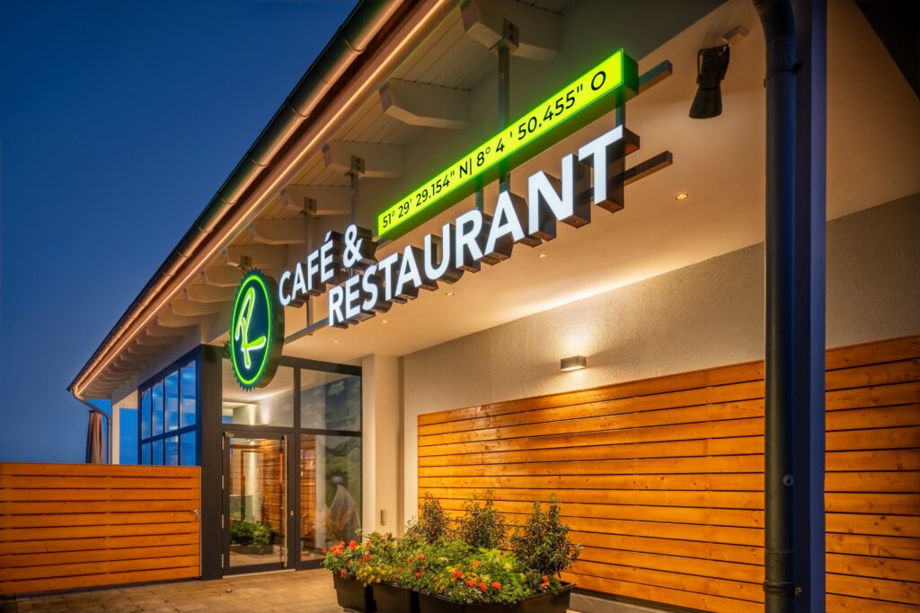 Upplyst Café & Restaurant-entré kvällstid med modern träfasad, glaspartier och blomlådor.