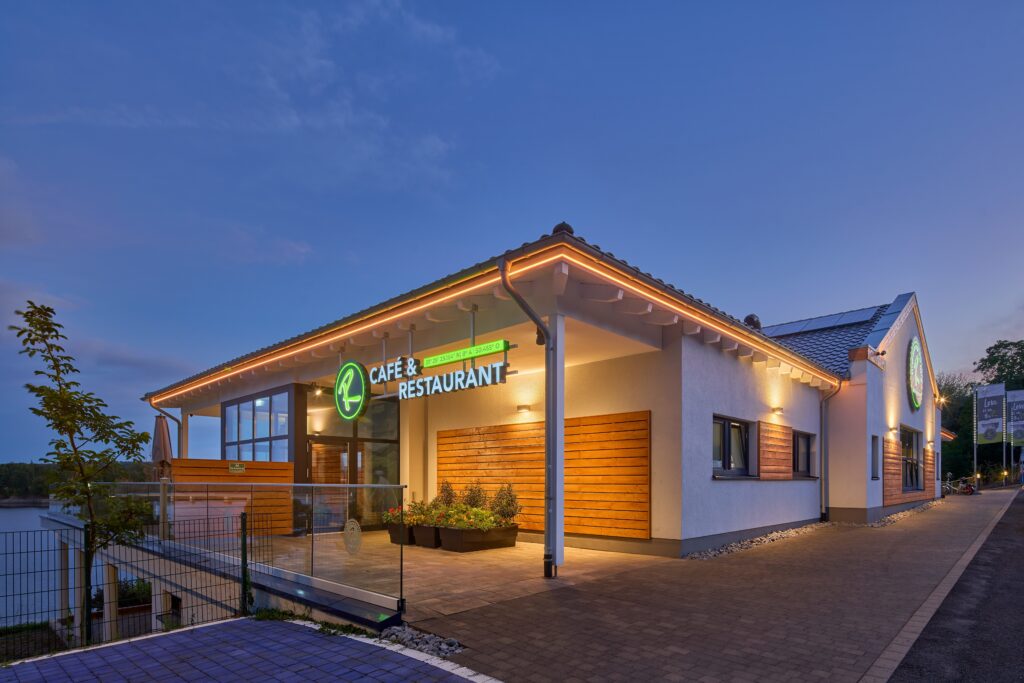 Modernt café och restaurang vid sjön i skymning, upplyst fasad och glasad uteservering.