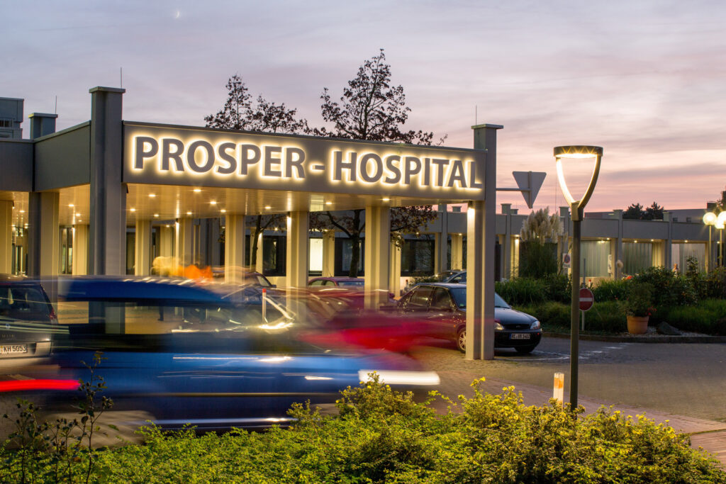 Entré till Prosper‑Hospital i skymning med varmvitt LED‑ljus från takarmaturer, skyltbelysning och en stilren pollare som effektivt lyser upp gångvägar och parkering och visar PROLJUS ABs energieffektiva och eleganta ljuslösningar som ökar trygghet och synlighet för personal och besökare