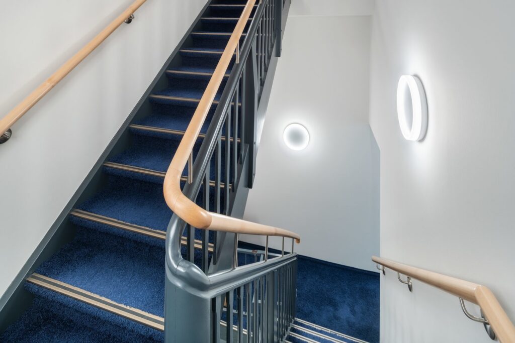 Trapphus med mörkblå matta där två runda väggarmaturer sprider mjukt, jämnt LED-ljus med modern ringdesign från PROLJUS AB som förbättrar säkerhet, estetik och energieffektivitet i trapphus och korridorer