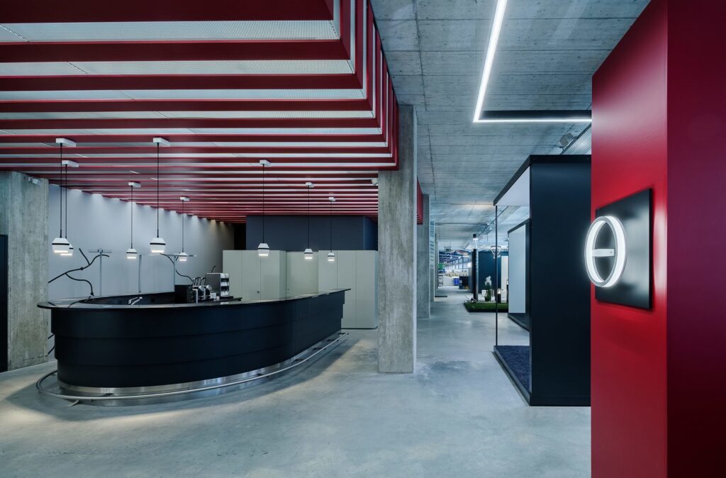 Modern kontorsinteriör med röda taklameller, stilrena vita pendellampor och kantade LED-linjer som belyser disk och gångstråk och visar PROLJUS ABs energieffektiva designarmaturer