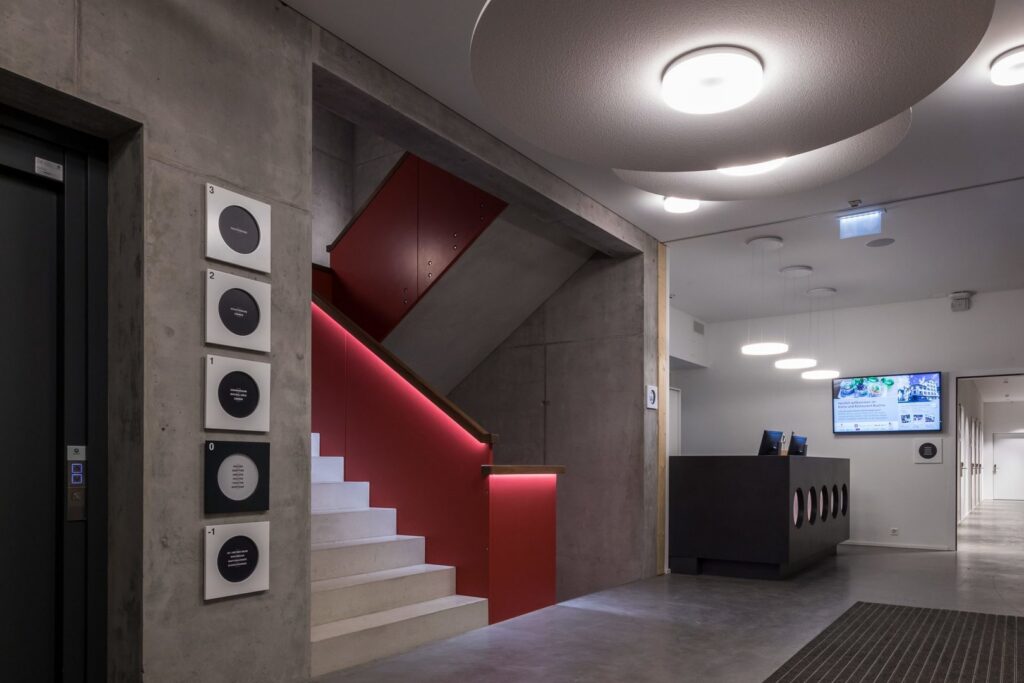 Entré med stora cirkulära takarmaturer och hängande diskpendlar samt LED-lister längs röd trappa, energieffektiva och dimbara armaturer från PROLJUS AB som skapar varmt, jämnt allmänljus och effektfull accentbelysning, kontakta PROLJUS AB för skräddarsydda belysningslösningar