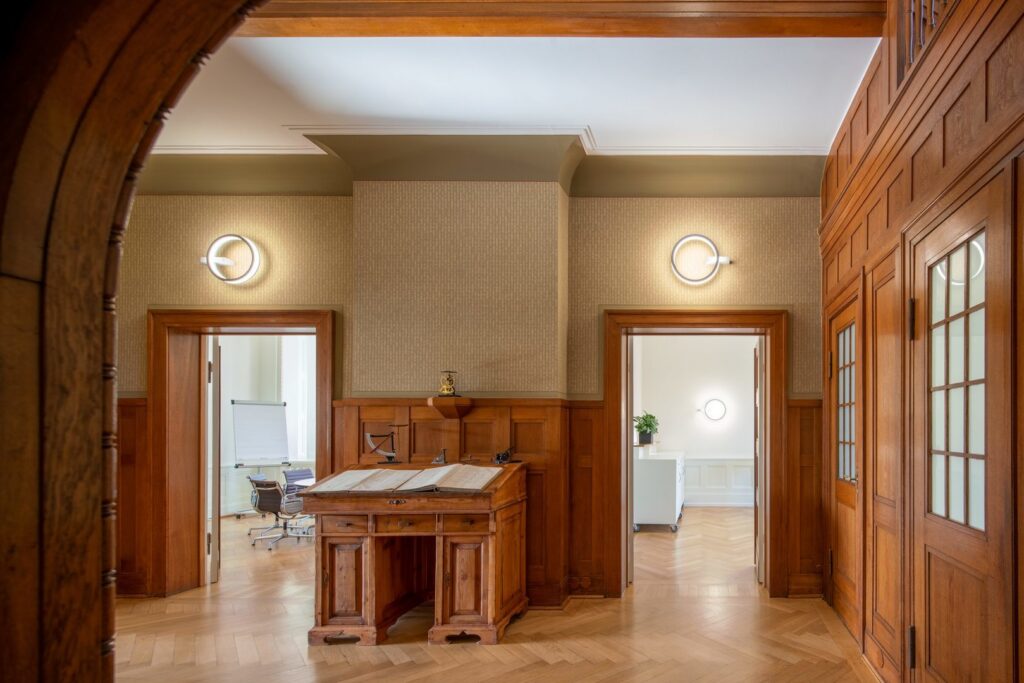 Träpanelhall med tre cirkulära väggarmaturer från PROLJUS AB som ger varm, jämn LED-belysning runt dörröppningar och skapar modern kontrast till klassisk inredning, energieffektiva armaturer perfekta för hotell, kontor och offentliga miljöer